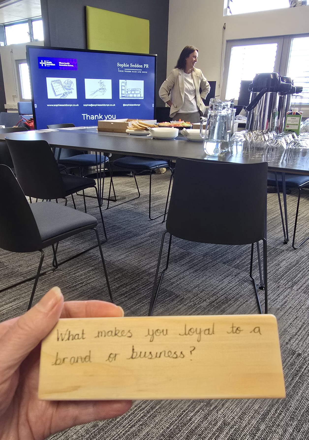 Person holding up a wooden Jenga block with the words “What makes you loyal to a brand or business?” written on it, while Sophie Seddon speaks in the background during a HHII workshop at The Glass Box, Huddersfield.